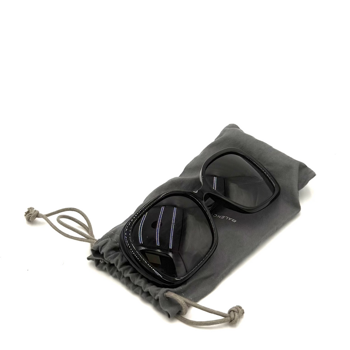 Black square-frame sunglasses with dark lenses resting on a gray drawstring pouch against a white background.