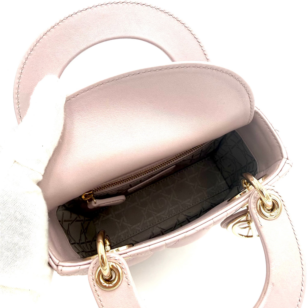 A light pink leather handbag shown open, revealing a dark patterned interior lining and gold-tone hardware. A white gloved hand is holding the bag against a plain white background.