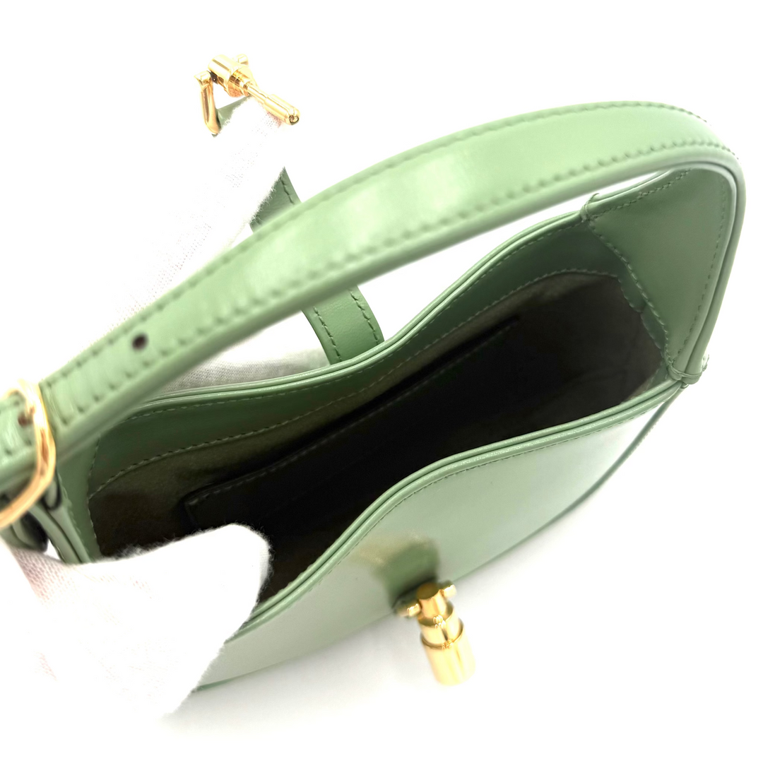 A sage green leather handbag shown open, revealing a black interior lining and gold-tone hardware, photographed on a white background with a white glove partially visible.