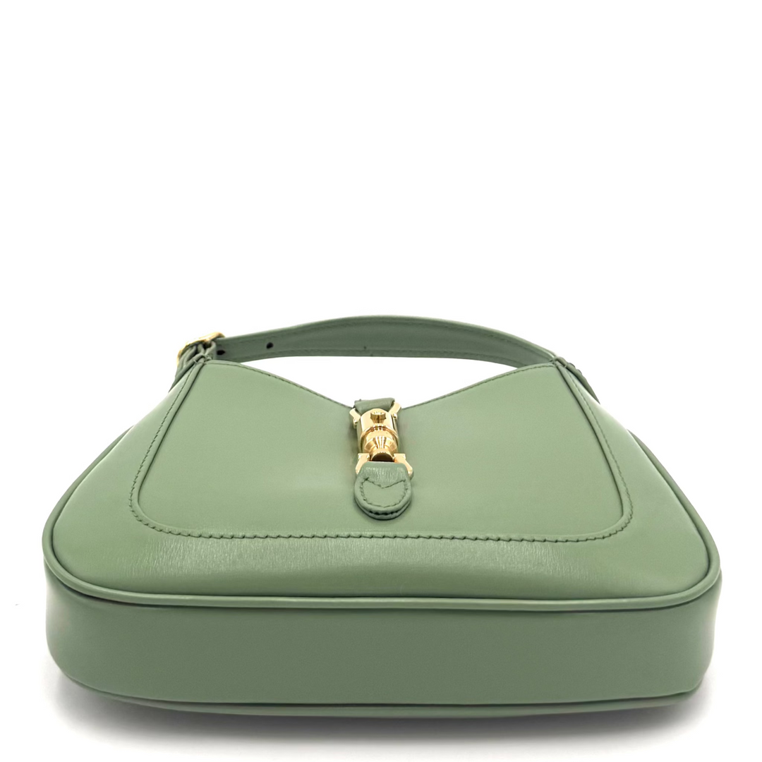A sage green leather shoulder bag with a structured, rounded triangular shape, featuring a gold-tone turn-lock clasp on the front flap and a matching green leather handle.