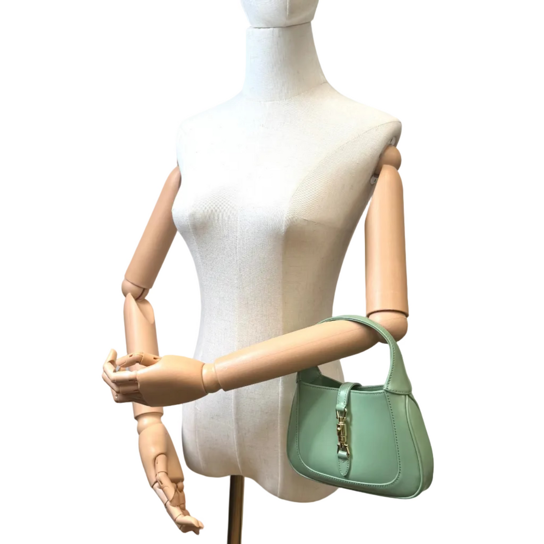 A mint green leather handbag with gold hardware, displayed on a white mannequin torso with articulated wooden arms against a white background.