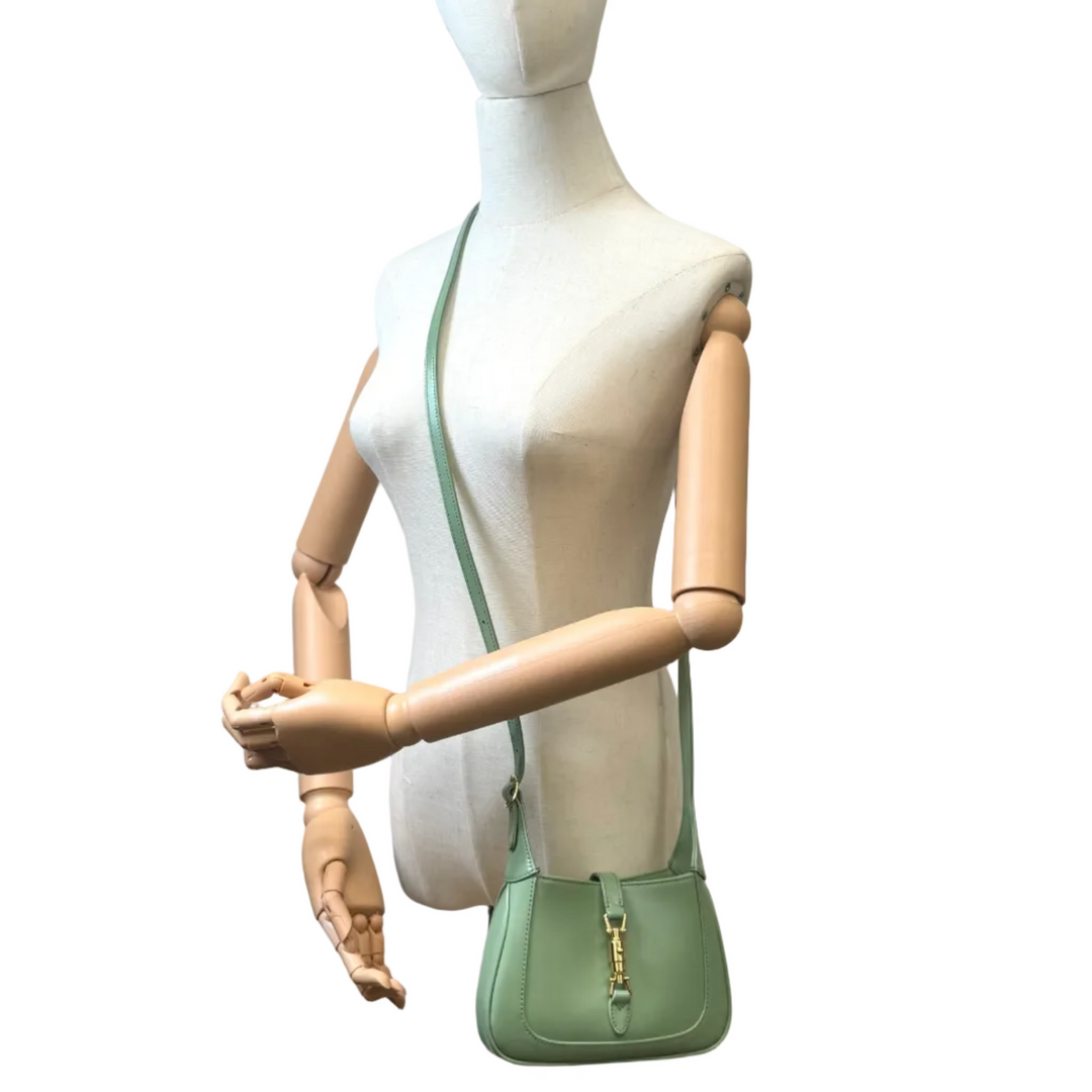 A sage green leather shoulder bag with gold-tone hardware, displayed on a white mannequin with wooden arms against a white background.