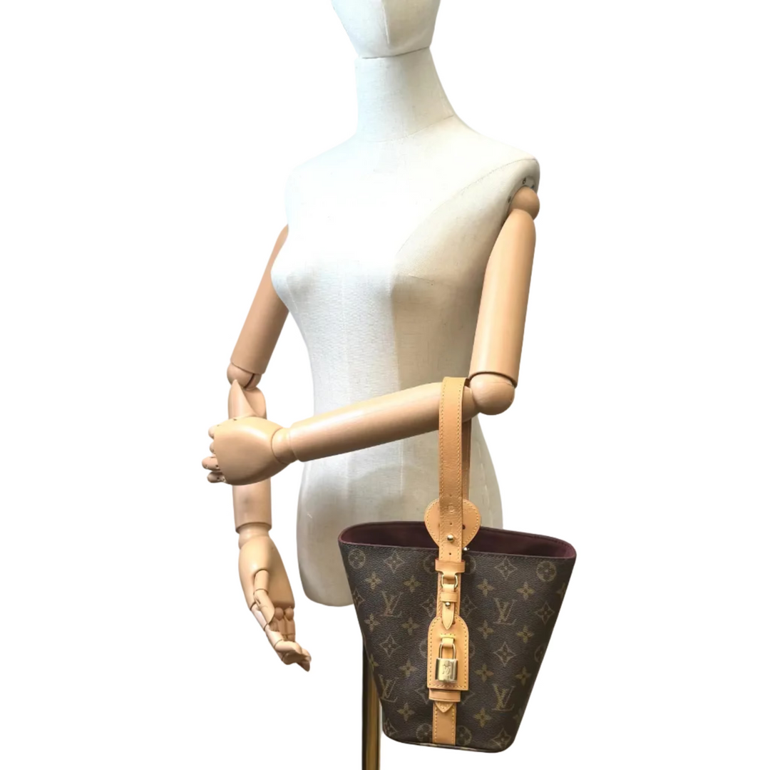 A Louis Vuitton monogram bucket bag with tan leather trim and a gold-tone buckle, displayed on a white mannequin torso with articulated wooden arms against a plain white background.