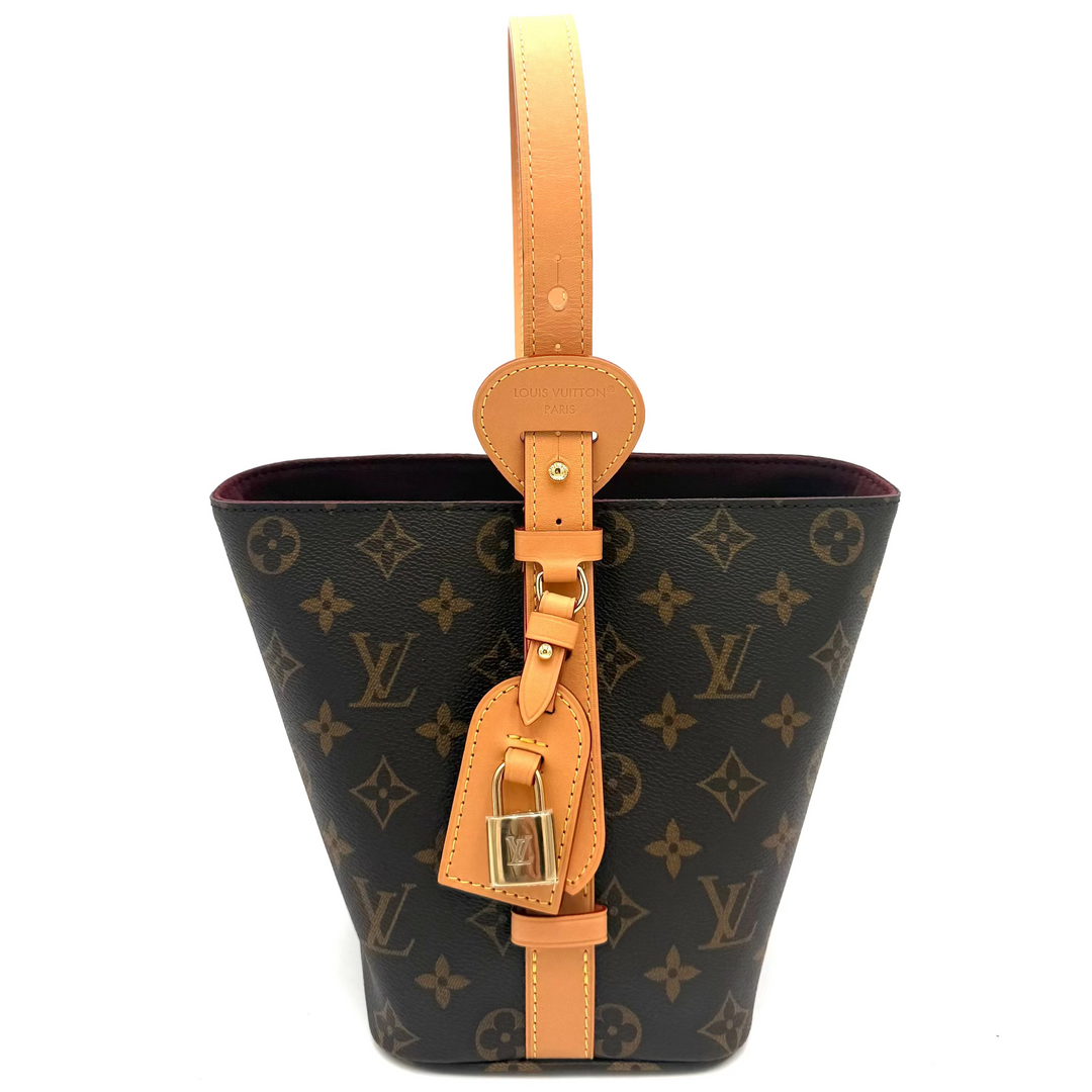 Louis Vuitton Monogram Canvas bucket bag with tan leather trim and handle, featuring gold-tone hardware and a hanging leather tag, shown against a plain white background.