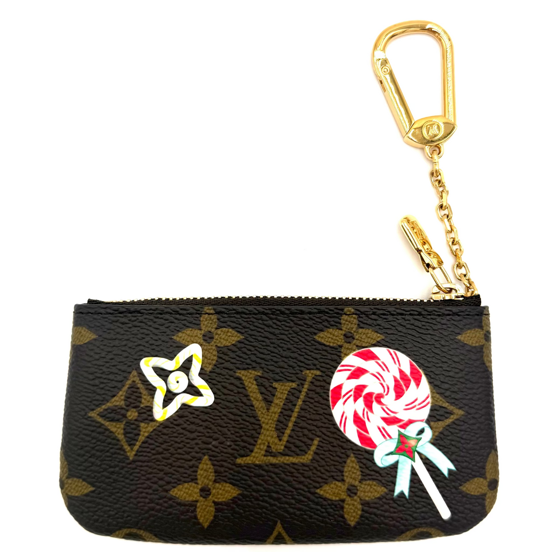 A Louis Vuitton monogram key pouch in brown coated canvas with gold-tone hardware, featuring a white and red lollipop charm and a yellow star flower charm.