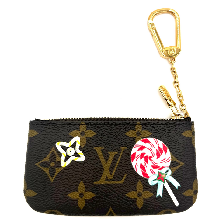 A Louis Vuitton monogram key pouch in brown coated canvas with gold-tone hardware, featuring a white and red lollipop charm and a yellow star flower charm.