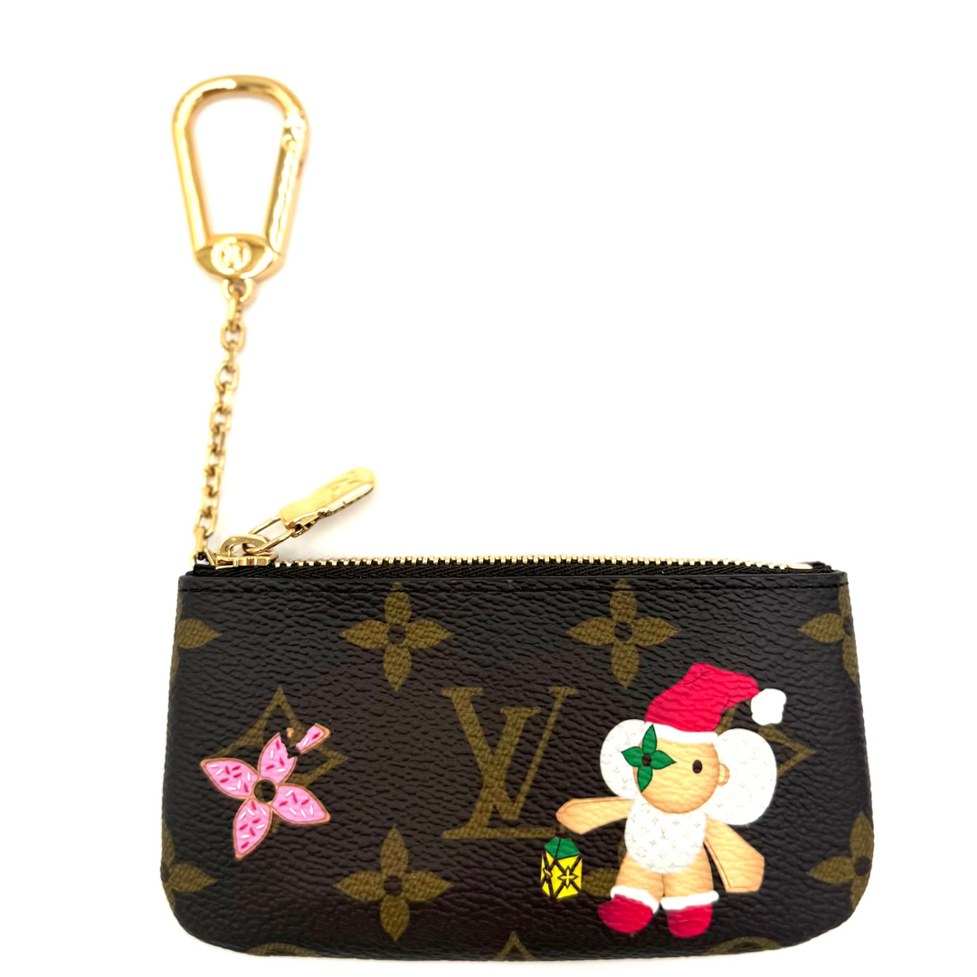 A Louis Vuitton monogram canvas key pouch with a gold-tone carabiner and chain, decorated with a pink flower patch and a Christmas-themed teddy bear applique.