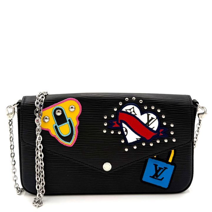 A black Louis Vuitton shoulder bag with a textured envelope flap, featuring three colorful patches (a yellow smiley face, a red-and-white heart with a red ribbon, and a blue LV patch) and silver studs, shown on a white background.
