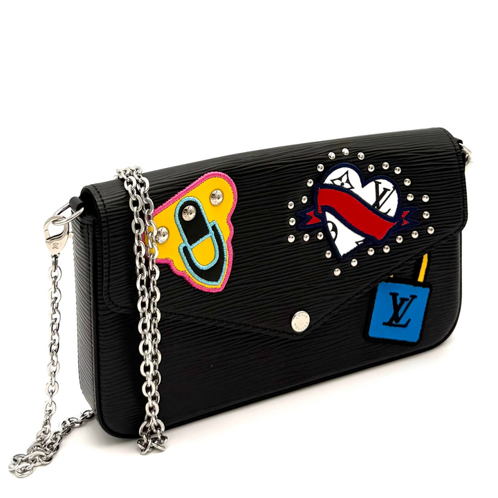 A black leather shoulder bag featuring multiple colorful patches, including a yellow and pink face patch, a red and white heart patch, and a blue Louis Vuitton monogram patch, with a silver chain strap.