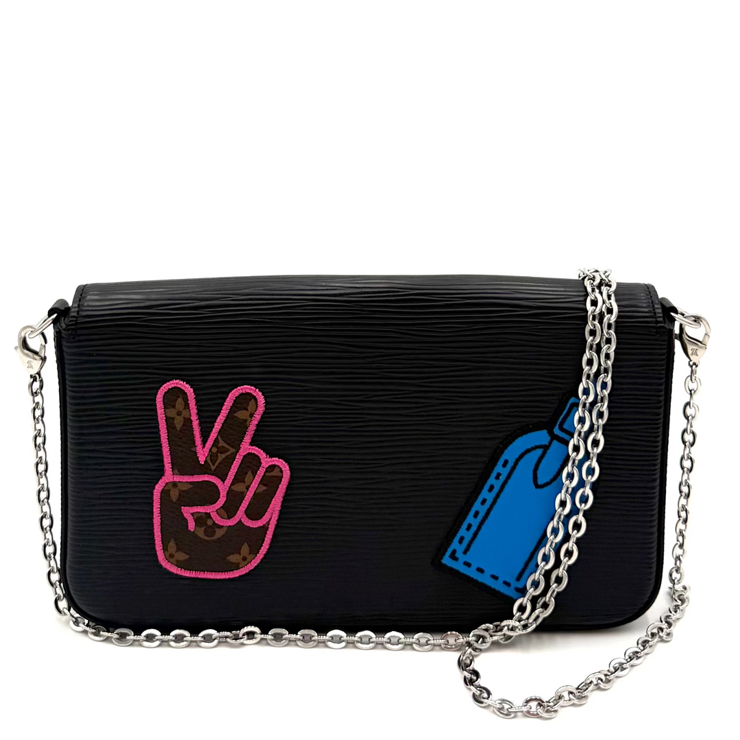 A black textured leather shoulder bag with a silver chain strap, featuring a pink peace sign patch and a blue price tag patch on the front.