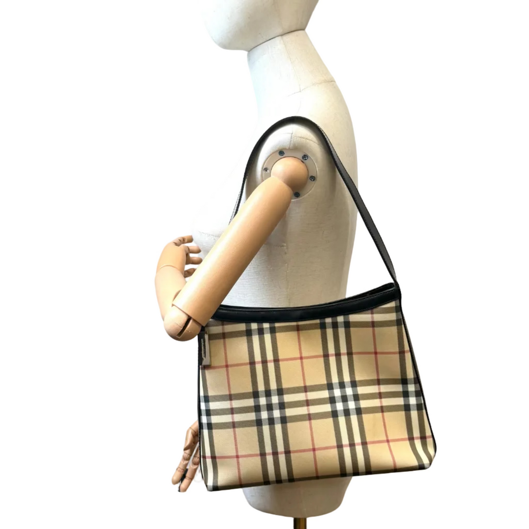 A beige plaid shoulder bag with black trim is displayed on a white mannequin torso in an indoor room with a white wall, painting, and wooden floor.