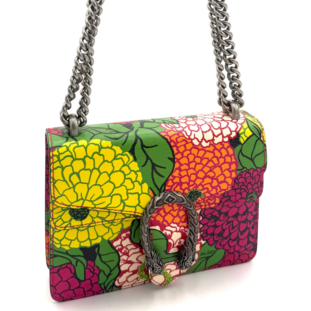 A small shoulder bag with a vibrant floral print featuring yellow, orange, pink, and green flowers, finished with a silver-tone chain strap and a central metal D-ring clasp.