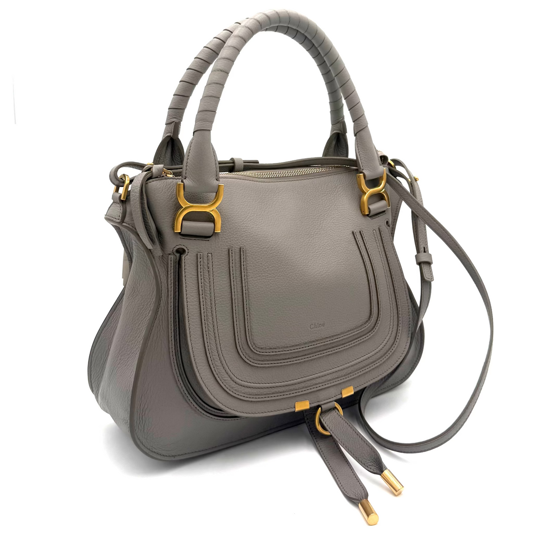 A gray leather handbag with structured double top handles, a front flap pocket, and gold-tone hardware, photographed against a white studio backdrop with visible softbox edges.