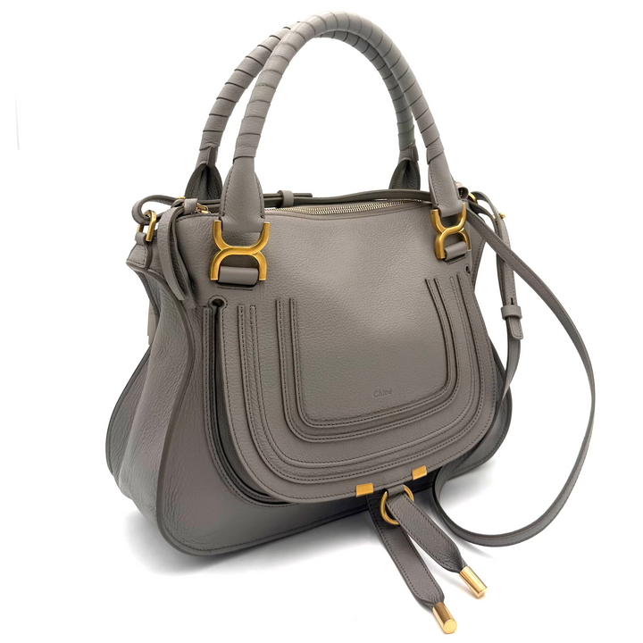 A gray leather handbag with structured double top handles, a front flap pocket, and gold-tone hardware, photographed against a white studio backdrop with visible softbox edges.