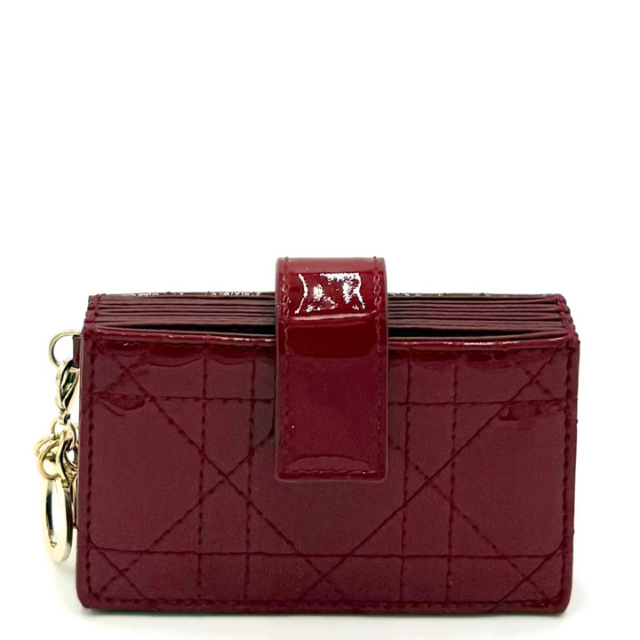 A deep burgundy patent leather card holder wallet with a quilted diagonal pattern and a matching strap closure featuring a silver-tone 'D' logo, shown on a white background with a gold keyring.