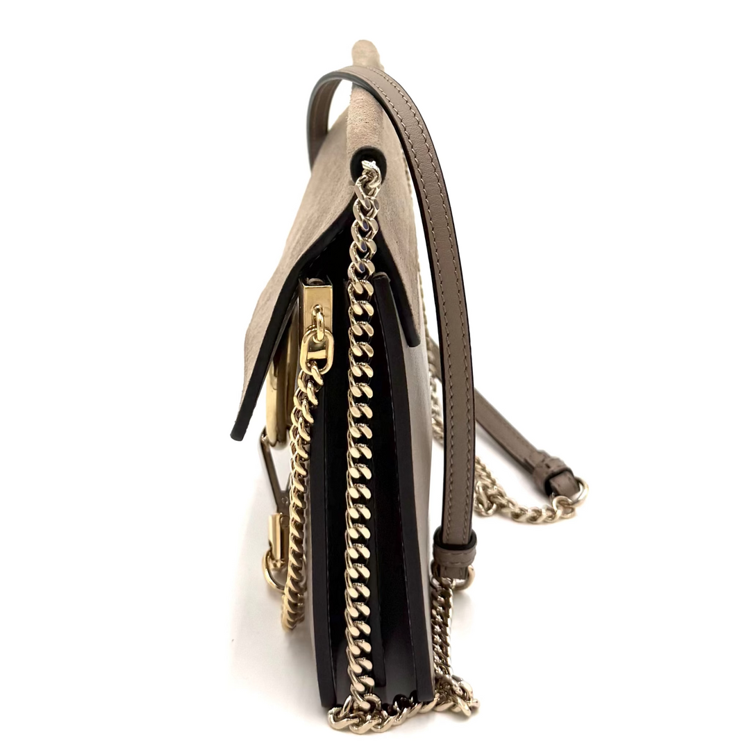 A small structured handbag shown from a side profile view on a plain white background, featuring taupe leather, a black leather panel, and gold-tone chain and zipper details.