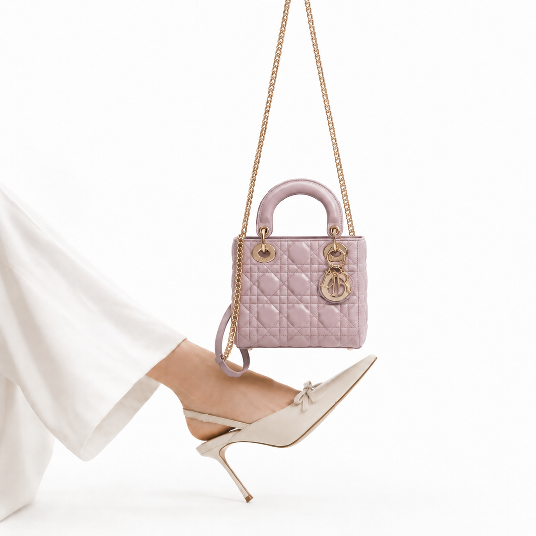 A light pink quilted Lady Dior handbag with gold-tone chain straps and a circular Dior logo charm, shown hanging against a white background with a model's foot in a white pointed-toe stiletto shoe.
