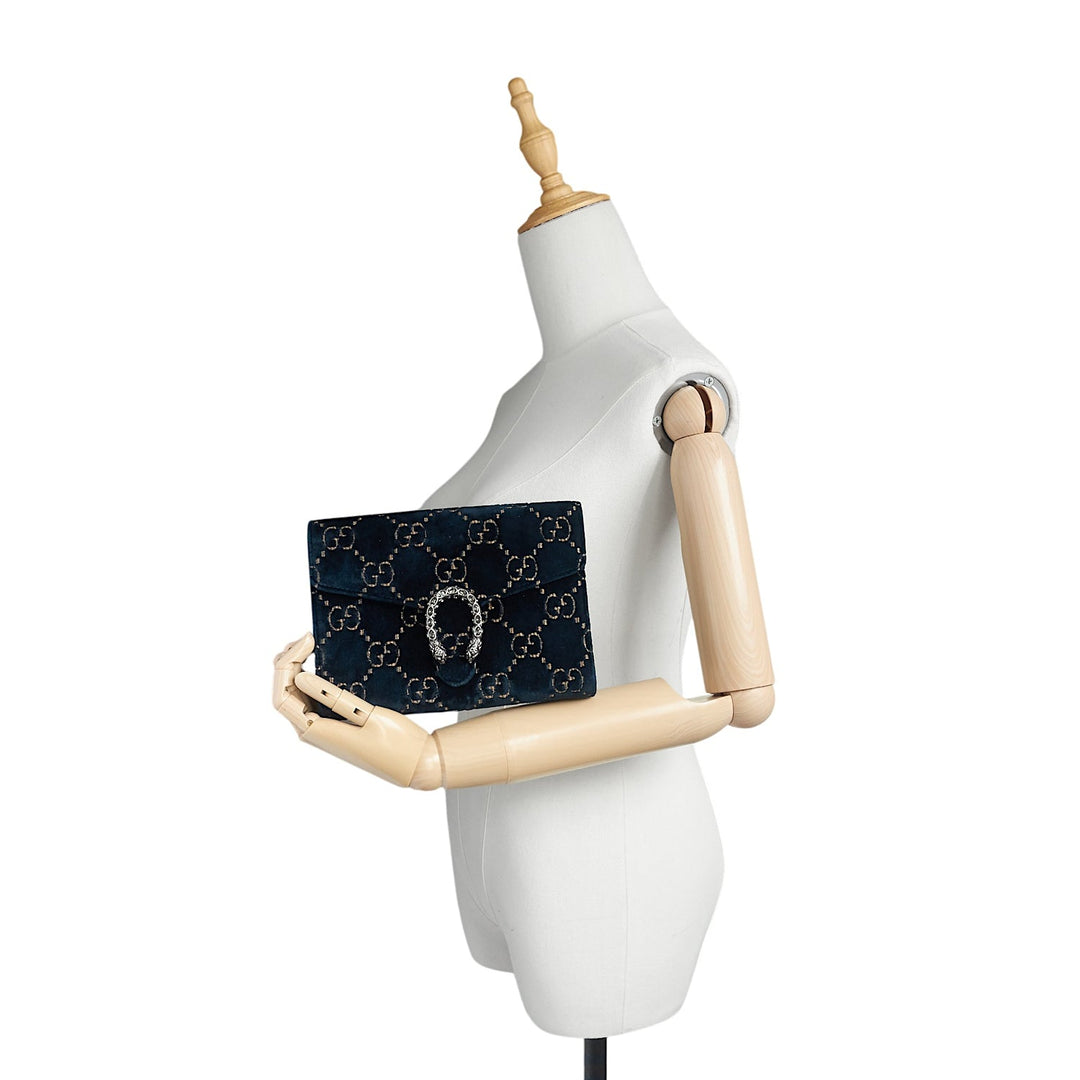 A navy blue Gucci clutch bag with a repeating GG monogram pattern and a central horsebit detail, held by a white wooden mannequin torso against a white background.