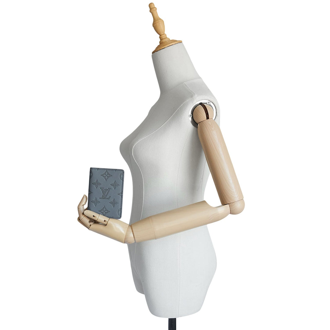 A Louis Vuitton monogrammed blue leather passport cover or small wallet held by a white mannequin torso with wooden arms on a white background.