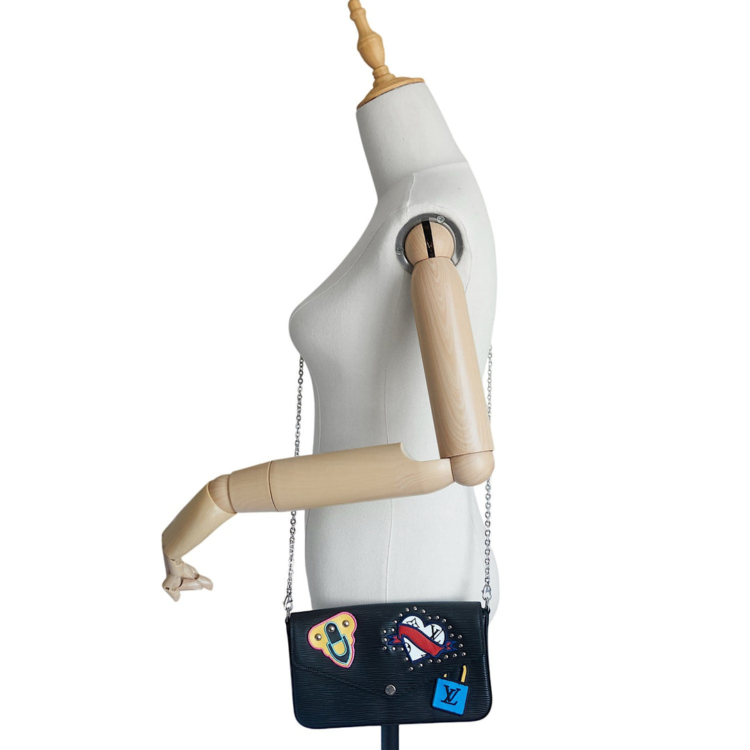 A black Louis Vuitton shoulder bag with colorful embroidered patches, displayed on a white mannequin torso with wooden arms against a white background.