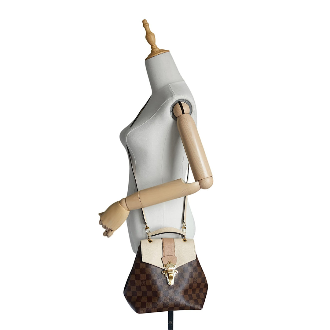 A Louis Vuitton shoulder bag in brown Damier canvas with a beige leather flap and trim, displayed on a white mannequin torso with wooden arms.